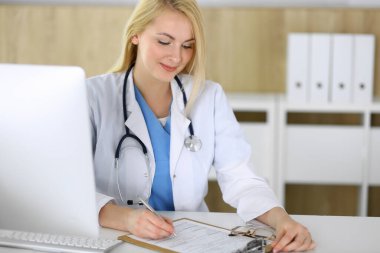 Hastanedeki ya da klinikteki masada oturan doktor kadın. Sarışın, neşeli doktor tıbbi kayıtları dolduruyor. Veriler ve tıp ve sağlık hizmetlerindeki en iyi hizmet