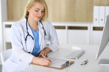 Hastanedeki ya da klinikteki masada oturan doktor kadın. Sarışın, neşeli doktor tıbbi kayıtları dolduruyor. Veriler ve tıp ve sağlık hizmetlerindeki en iyi hizmet
