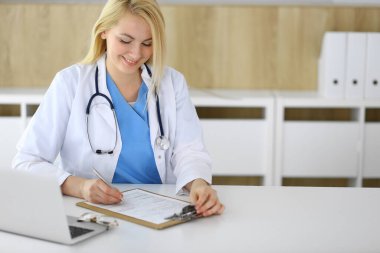 Hastanedeki ya da klinikteki masada oturan doktor kadın. Sarışın, neşeli doktor tıbbi kayıtları dolduruyor. Veriler ve tıp ve sağlık hizmetlerindeki en iyi hizmet