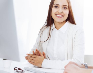 Genç, neşeli bir kadın bilgisayarla masada oturuyor ve güneşli ofisteki kameraya bakıyor. Öğrenci kız ya da iş kadınına benziyor. Vesikalık