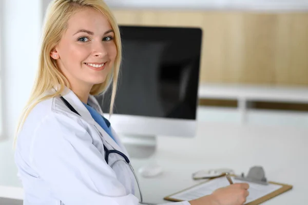 Läkare kvinna på jobbet medan du sitter vid skrivbordet på sjukhus eller klinik. Blond glad läkare redo att hjälpa patienter. Data och bästa service inom medicin och sjukvård — Stockfoto
