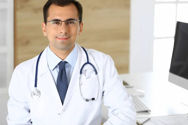 Doutor homem sentado na mesa em seu local de trabalho e sorrindo para a câmera. Serviço médico perfeito na clínica. Futuro feliz da medicina e da saúde — Fotografia de Stock