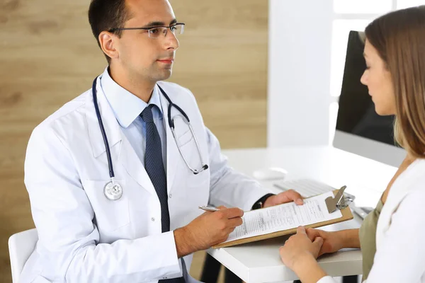 Doctor and patient discussing the results of a physical examination while sitting at a desk in a clinic. A male doctor using a clipboard to fill out a medical history of a young womans medication