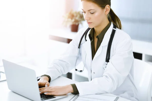 책상에 앉아서 맑은 병원 사무실에서 노트북으로 타이핑하는 여자 - 의사. 의사 가일하고 있다 — 스톡 사진