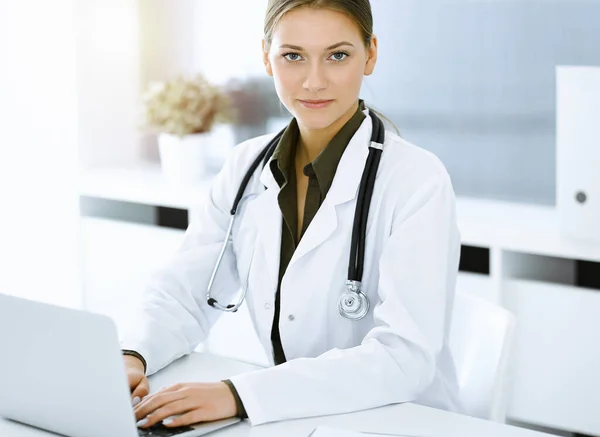 Mulher-médico digitando no computador portátil enquanto sentado na mesa no escritório do hospital ensolarado. Médico a trabalhar — Fotografia de Stock