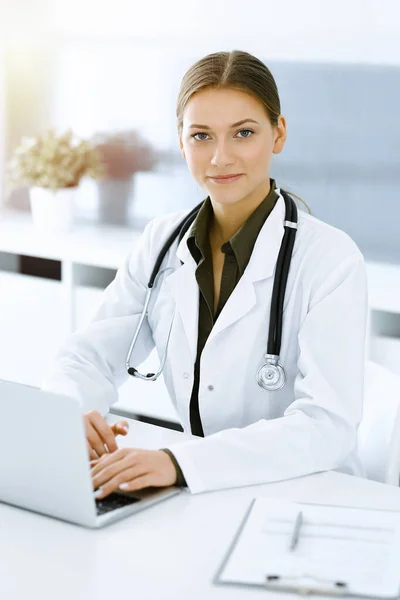 Woman-doctor typing on laptop computer while sitting at the desk in sunny hospital office. Physician at work