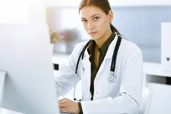 Mujer-médico escribiendo en la computadora PC mientras está sentado en el escritorio en la oficina del hospital soleado. Datos en medicina y asistencia sanitaria — Foto de Stock