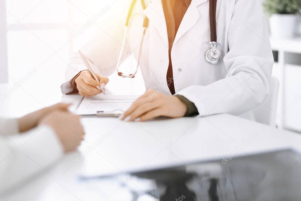 Unknown woman-doctor and female patient sitting and talking at medical examination in sunny clinic, close-up. Therapist filling up medication history record. Medicine concept