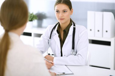 Kadın doktor ve hasta hastane muayenehanesinde oturup konuşuyorlar. Yeşil renkli bluz terapiste yakışır. Tıp ve sağlık hizmetleri kavramı