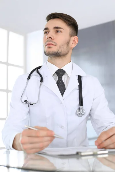 Doctor hombre llenando el formulario de historial de medicamentos mientras está sentado en el escritorio de cristal en el consultorio médico o clínica. Concepto de medicina y salud — Foto de Stock