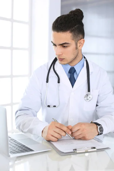 在医务室或诊所的玻璃桌上，阿拉伯医生在填写药物病历记录时使用笔记本电脑。医药和保健概念 — 图库照片