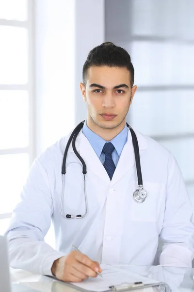 Homem médico árabe usando computador portátil durante o preenchimento de registros de histórico de medicação na mesa de vidro no consultório médico ou clínica. Conceito de medicina e saúde — Fotografia de Stock