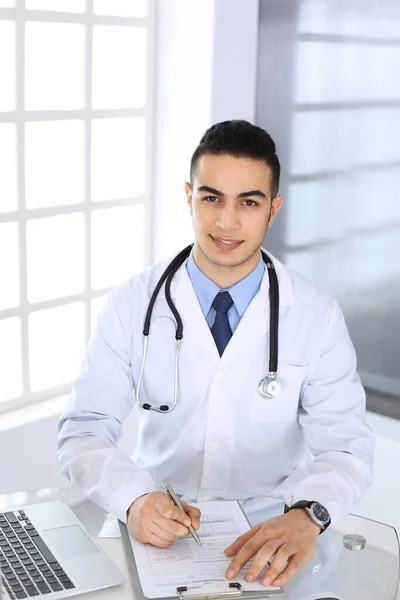 Homem médico árabe usando computador portátil durante o preenchimento de registros de histórico de medicação na mesa de vidro no consultório médico ou clínica. Conceito de medicina e saúde — Fotografia de Stock