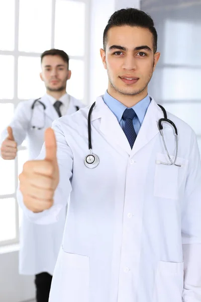 Άραβας γιατρός δείχνει Ok υπογράψει με τους αντίχειρες επάνω με καυκάσιος συνάδελφος στο ιατρικό γραφείο ή κλινική. Διαφορετική ομάδα γιατρών, καλύτερη θεραπεία, ιατρική και υγειονομική περίθαλψη έννοια — Φωτογραφία Αρχείου