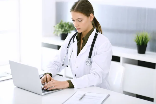 Médico digitando no computador portátil enquanto sentado na mesa no escritório do hospital. Médica no trabalho. Dados em medicina e cuidados de saúde — Fotografia de Stock