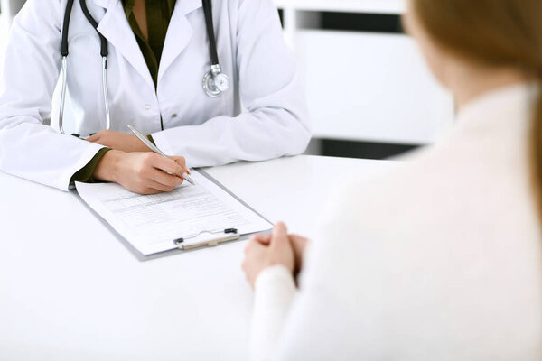 Woman doctor and patient sitting and talking at medical examination at hospital office, close-up. Therapist filling up medication history records. Medicine and healthcare concept