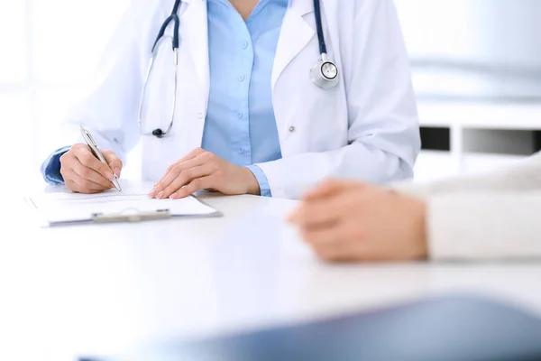 Doctora y paciente sentadas y hablando en el examen médico en el consultorio del hospital, de cerca. Médico llenando registros de historial de medicamentos. Concepto de medicina y salud — Foto de Stock