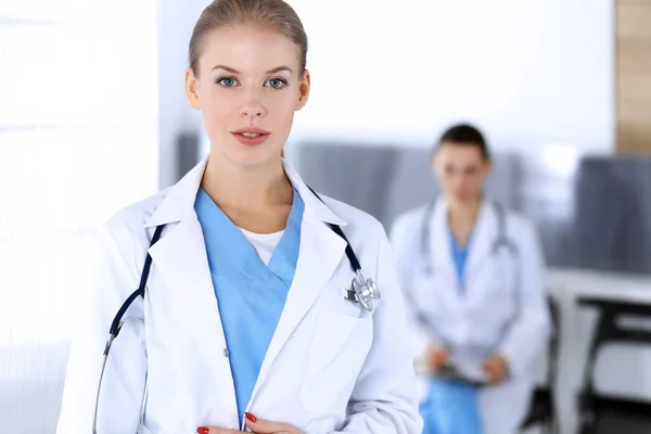 Doctora mujer de pie en la oficina del hospital de emergencia con su colega de fondo. Médico en el trabajo, retrato de estudio. Concepto de medicina y salud — Foto de Stock