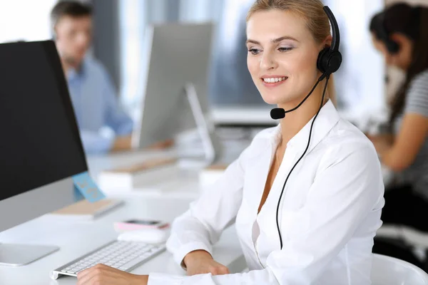 Blonde business woman using headset for communication and consulting people at customer service office. Call center. Group of operators at work at the background — Stock Photo, Image