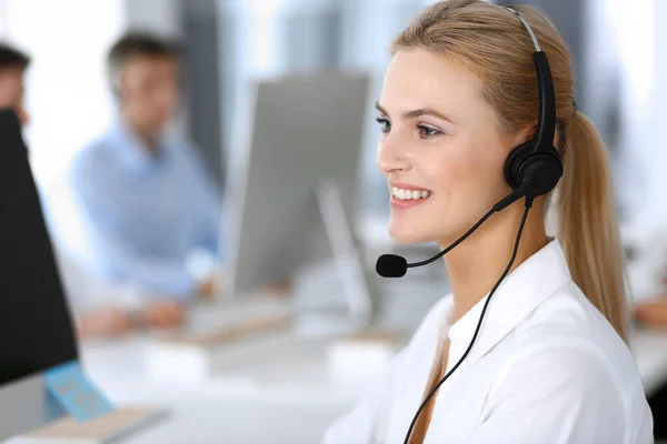 Blonde business woman using headset for communication and consulting people at customer service office. Call center. Group of operators at work at the background — Stock Photo, Image