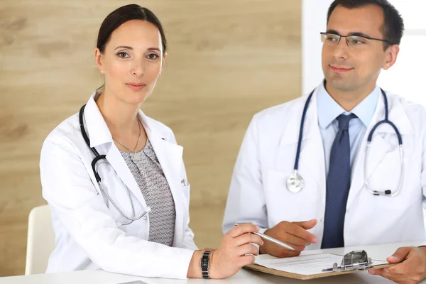 Middle aged woman doctor filling up medical documents or prescription with her male colleague. Group of doctors at work discussing treatment problems while sitting at a desk in hospital. Data in