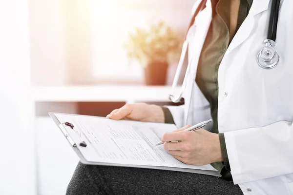 Mujer médica desconocida escribiendo algo en el portapapeles mientras está sentada en una clínica soleada, de cerca. El terapeuta que usa blusa verde en el trabajo está llenando el historial de medicamentos. Concepto de medicina — Foto de Stock