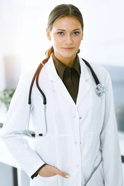 Mujer-médico de pie y mirando a la cámara en la clínica soleada. Un servicio médico perfecto. Medicina y asistencia sanitaria —  Fotos de Stock
