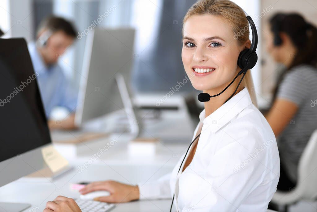 Blonde business woman using headset for communication and consulting people at customer service office. Call center. Group of operators at work at the background