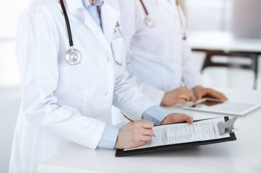 İş yerindeki bilinmeyen kadın doktorlar, arka planda hastaları var. Güneşli hastane resepsiyonunda beklerken tıbbi belgeleri dolduran kadın doktorlar, yakın plan. Sağlık hizmetleri