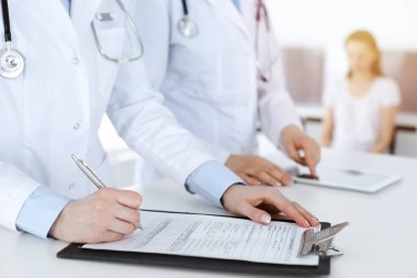 İş yerindeki bilinmeyen kadın doktorlar, arka planda hastaları var. Güneşli hastane resepsiyonunda beklerken tıbbi belgeleri dolduran kadın doktorlar, yakın plan. Sağlık hizmetleri