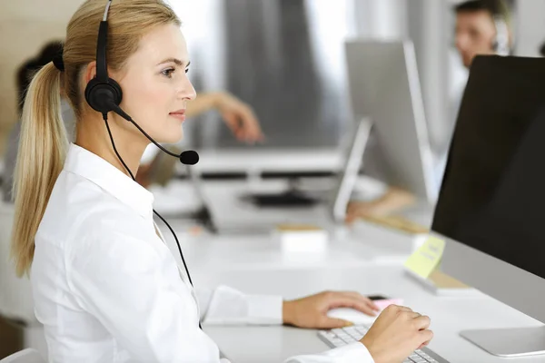 Blonde business woman using headset for communication and consulting people at customer service office. Call center. Group of operators at work — Stock Photo, Image