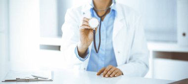 Bilinmeyen mavi bluzlu kadın doktor, klinikteki masada otururken steteskop başı tutuyor, yakın plan. Doktor hastayı muayene etmeye ve yardım etmeye hazır. Tıp konsepti