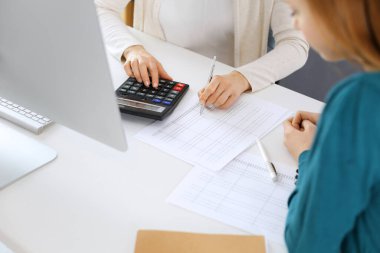 Muhasebeci mali durumu kontrol ediyor ya da vergi formları için hesap makinesi geliri ile sayıyor, eller yakın çekim. İş kadını ofisteki masada iş arkadaşıyla oturuyor ve çalışıyor. Denetim kavramı