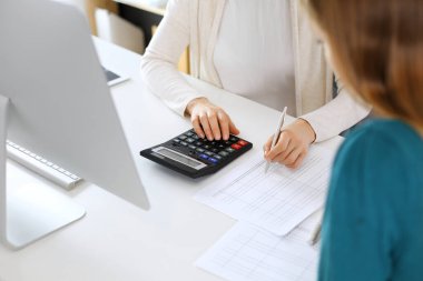 Muhasebeci mali durumu kontrol ediyor ya da vergi formları için hesap makinesi geliri ile sayıyor, eller yakın çekim. İş kadını ofisteki masada iş arkadaşıyla oturuyor ve çalışıyor. Denetim kavramı