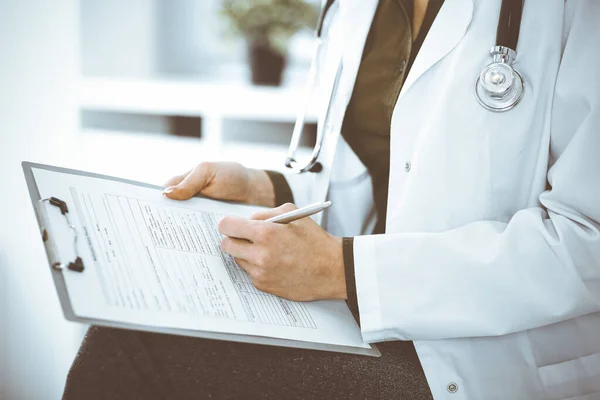 Mujer médica desconocida escribiendo algo en el portapapeles mientras está sentada en la silla, de cerca. El terapeuta que usa blusa verde en el trabajo está llenando el historial de medicamentos. Concepto de medicina — Foto de Stock