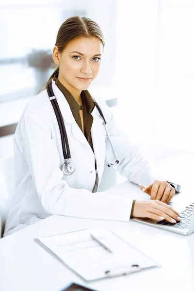 Woman-doctor typing on laptop computer while sitting at the desk in hospital office. Physician at work