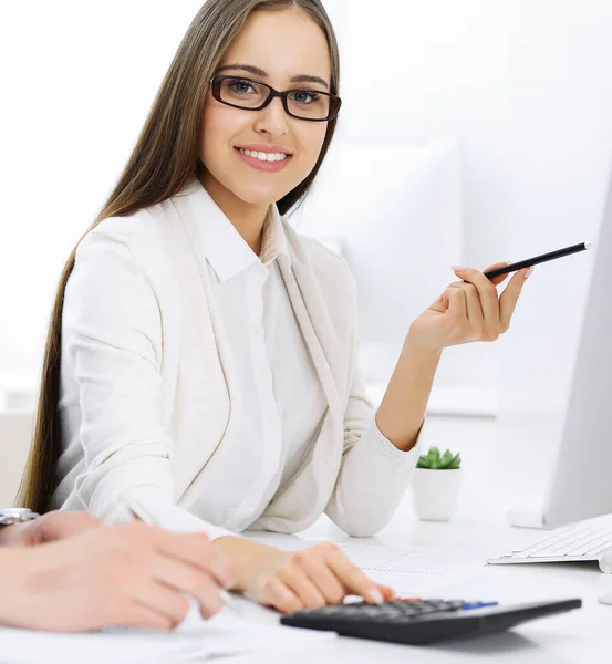 Young cheerful woman sitting at the desk with computer and looking at camera in white colored office. Looks like student girl or business lady. Headshot
