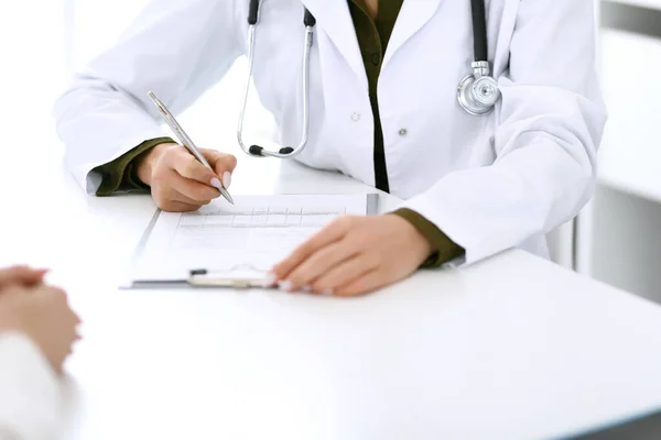 Doctora y paciente sentadas y hablando en el examen médico en el consultorio del hospital, de cerca. Terapeuta llenando los historiales de medicación. Concepto de medicina y salud — Foto de Stock