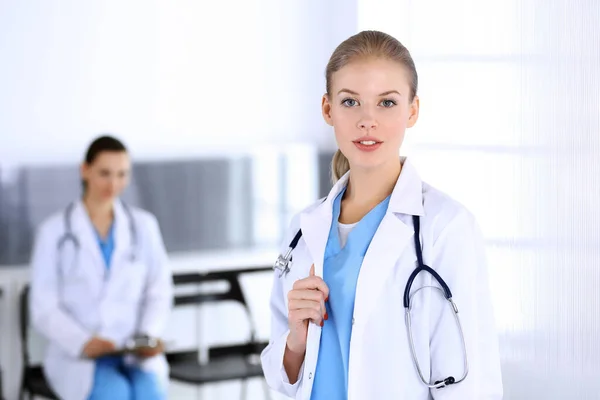 Orvos nő áll a sürgősségin a kollégájával a háttérben. Orvos a munkahelyén, műtermi portré. Orvosi és egészségügyi ellátás fogalma — Stock Fotó