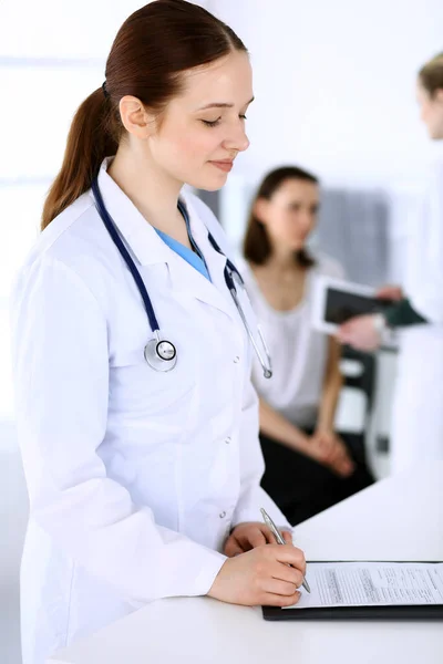 バックグラウンドで患者と同僚と仕事をしている医師の女性。病院の受付に立っている間、医師は医療文書や処方箋を記入します。医療・ヘルスケアのデータ — ストック写真