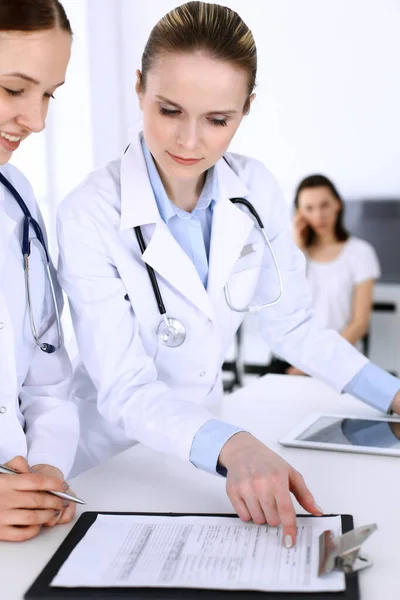 Group of doctors at work in hospital with patient in a queue at the background. Physician filling up medical documents or prescription while standing at reception desk. Data in medicine and health — Stock Photo, Image