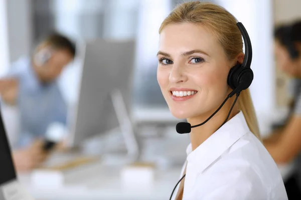 Mujer rubia de negocios con auriculares para la comunicación y la consulta de las personas en la oficina de servicio al cliente. Centro de llamadas. Grupo de operadores en el trabajo en segundo plano — Foto de Stock