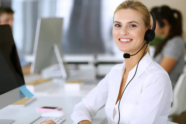 金发女商人使用耳机与客户服务办公室的人进行交流和咨询。呼叫中心背景下工作的操作员小组 — 图库照片