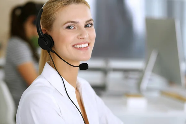 Blond affärskvinna använder headset för kommunikation och konsultera människor på kundtjänst kontor. Samtalscentralen. Grupp av aktörer i arbete i bakgrunden — Stockfoto
