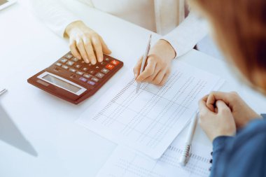 Muhasebeci mali durumu kontrol ediyor ya da vergi formları için hesap makinesi geliri ile sayıyor, eller yakın çekim. İş kadını ofisteki masada iş arkadaşıyla oturuyor ve çalışıyor. Denetim kavramı