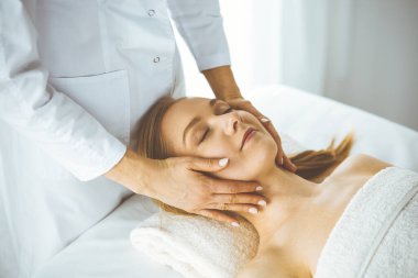 Güzel, mutlu bir kadın kaplıca salonunda kapalı gözlerle yüz masajından zevk alıyor. Tıp ve Güzellik konseptinde rahatlatıcı tedavi