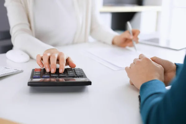 Účetní kontroluje finanční výkaz nebo počítá příjmy kalkulačky pro daňové formuláře, ruce detailní. Obchodní žena, která seděla a pracovala s kolegyní u psacího stolu v kanceláři, modře tónovala. Daň a — Stock fotografie