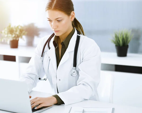 Mulher-médico digitando no computador portátil enquanto sentado na mesa no escritório do hospital ensolarado. Médico a trabalhar — Fotografia de Stock