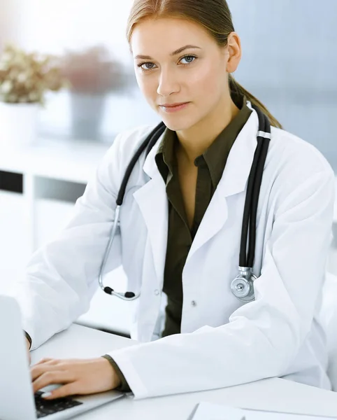 Woman-doctor typing on laptop computer while sitting at the desk in sunny hospital office. Physician at work