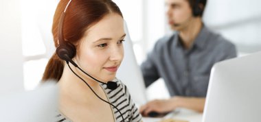 İnternette müşterilerle konuşurken kulaklık ve bilgisayar kullanan sıradan giyinmiş genç bir kadın. Güneşli ofiste çalışan bir grup operatör. Çağrı merkezi